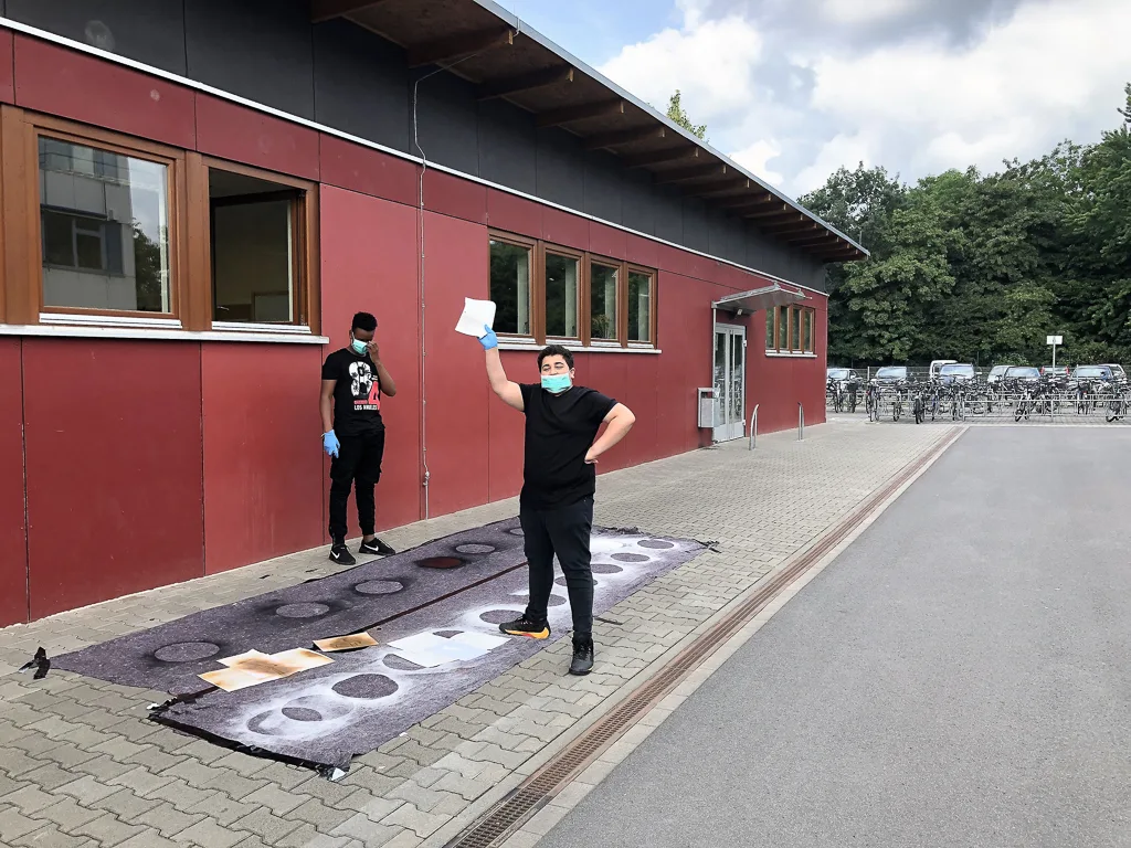 Zwei Personen stehen vor dem Schulgebäude auf einer Plane.